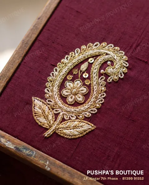 Close-up of hand aari embroidery: a gold and ivory paisley in raised chain stitch on maroon silk, with sequins and pearls sewn on one by one, still in its wooden frame