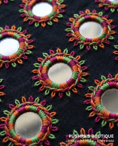 Close-up of hand mirror work on black cotton: round mirrors held by rings of magenta, orange and green stitches, with chain-stitch petals
