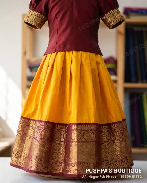 Girl's mango-yellow pattu pavadai with a wide maroon and gold zari border and a maroon blouse with zari puff sleeves